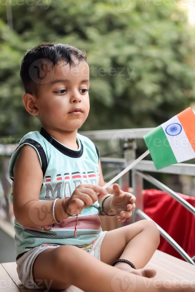 Cute Indian boy holding Indian National Flag in his hangs at home balcony. Bright portrait of happy child sitting on the table with national flag photo