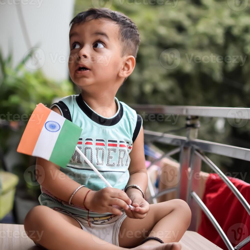 Cute Indian boy holding Indian National Flag in his hangs at home balcony. Bright portrait of happy child sitting on the table with national flag photo