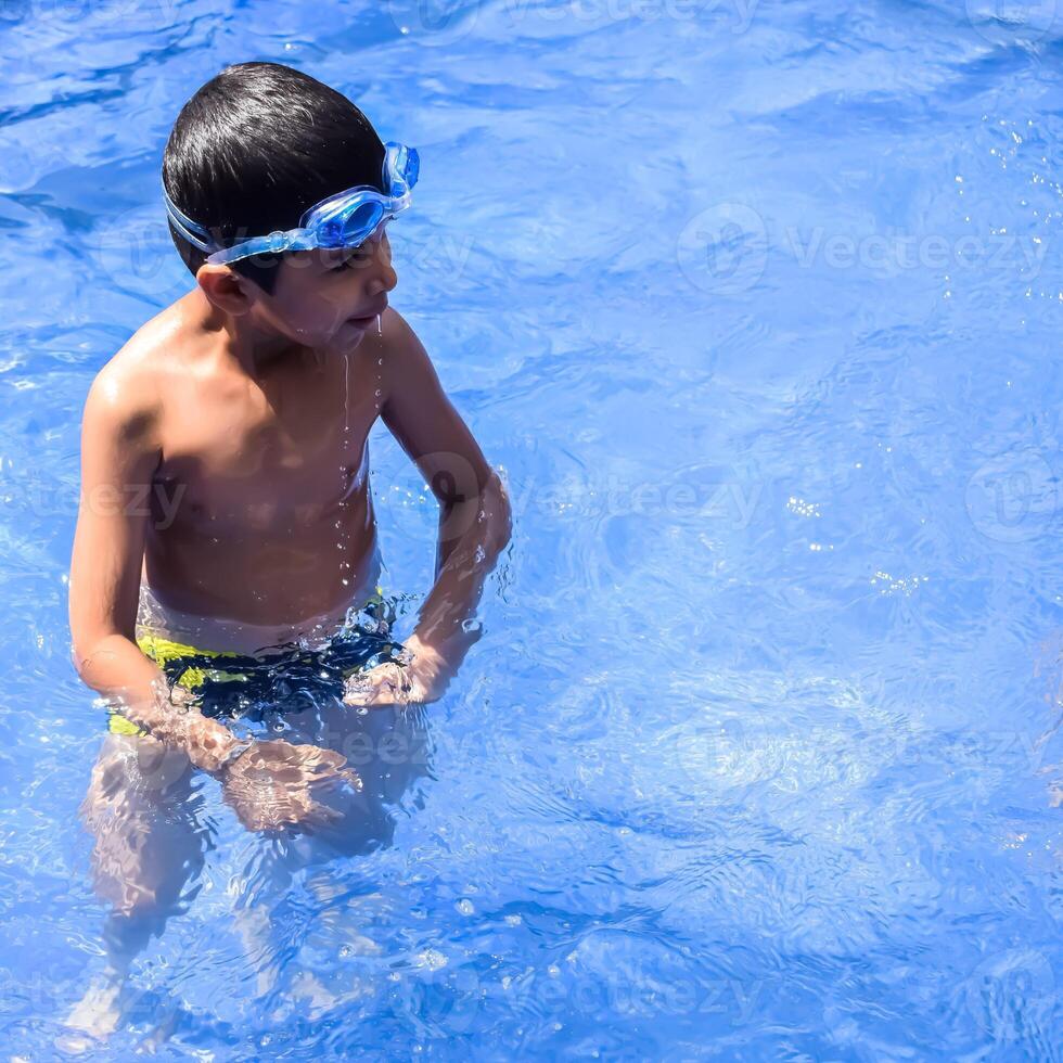 Happy Indian boy swimming in a pool, Kid wearing swimming costume along with air tube during hot summer vacations, Children boy in big swimming pool. photo
