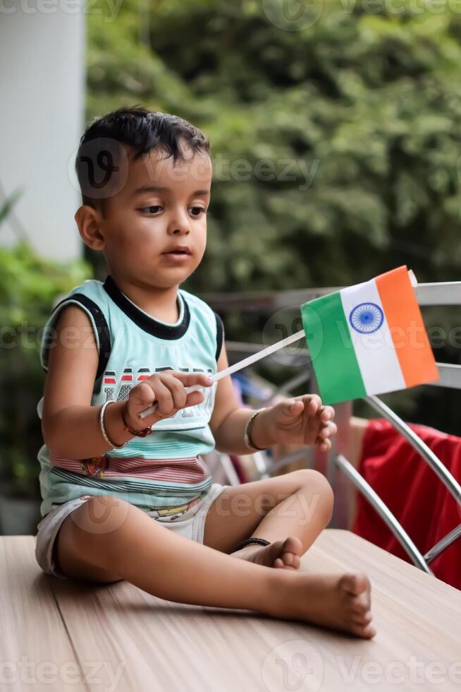 Cute Indian boy holding Indian National Flag in his hangs at home balcony. Bright portrait of happy child sitting on the table with national flag photo
