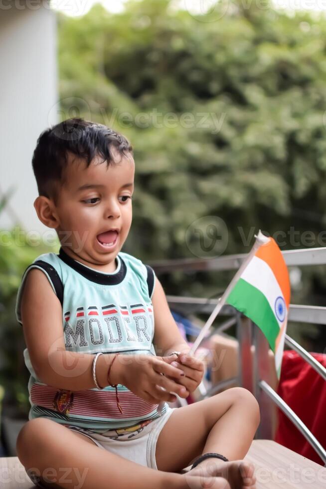 Cute Indian boy holding Indian National Flag in his hangs at home balcony. Bright portrait of happy child sitting on the table with national flag photo