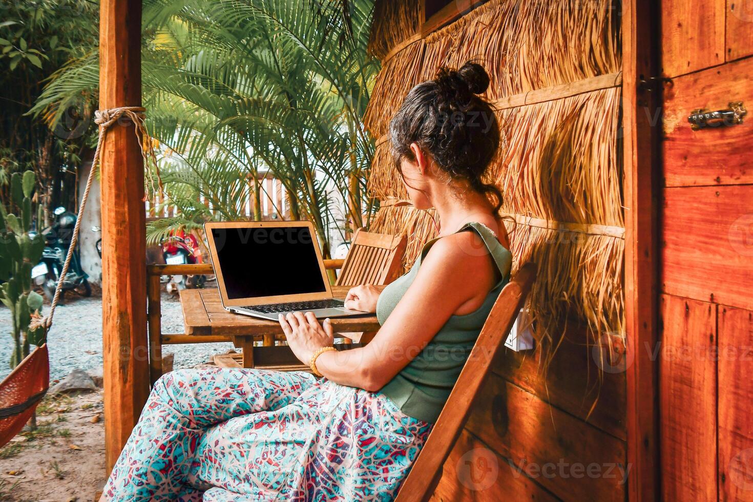 Back view caucasian hipster woman sit by computer outside eco-friendly straw house building guesthouse. Freelance work in south-east asia remotely isolated in tropical nature concept photo
