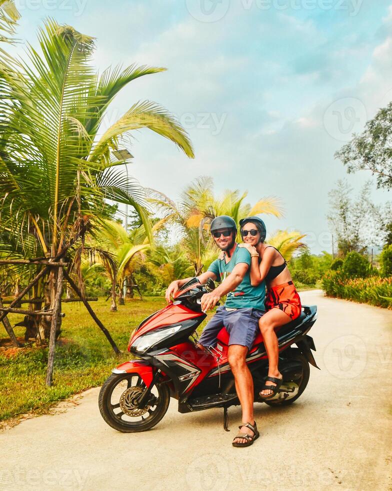 Happy smiling couple travelers riding motorbike scooter in safety helmets during tropical vacation under palm trees on Phu Quoc island , Vietnam photo