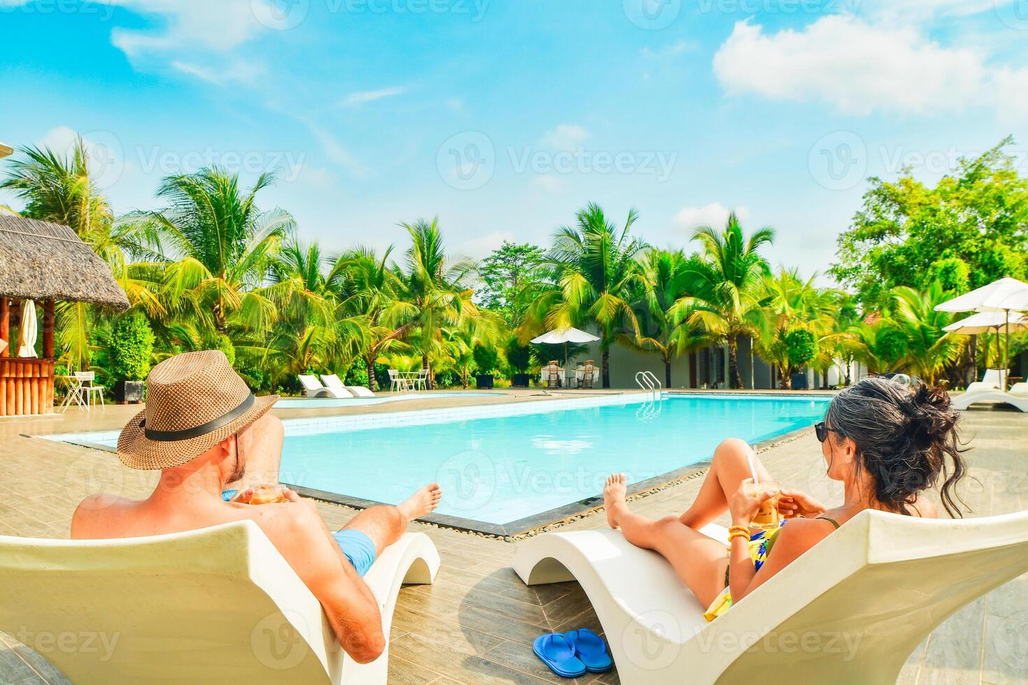 Couple lay on sunbeds enjoy sunbathing drink cold beverage on all-inclusive vacation holidays at tropical resort with swimming pool surrounded by coconut palm trees by coast on sunny hot tropical day photo