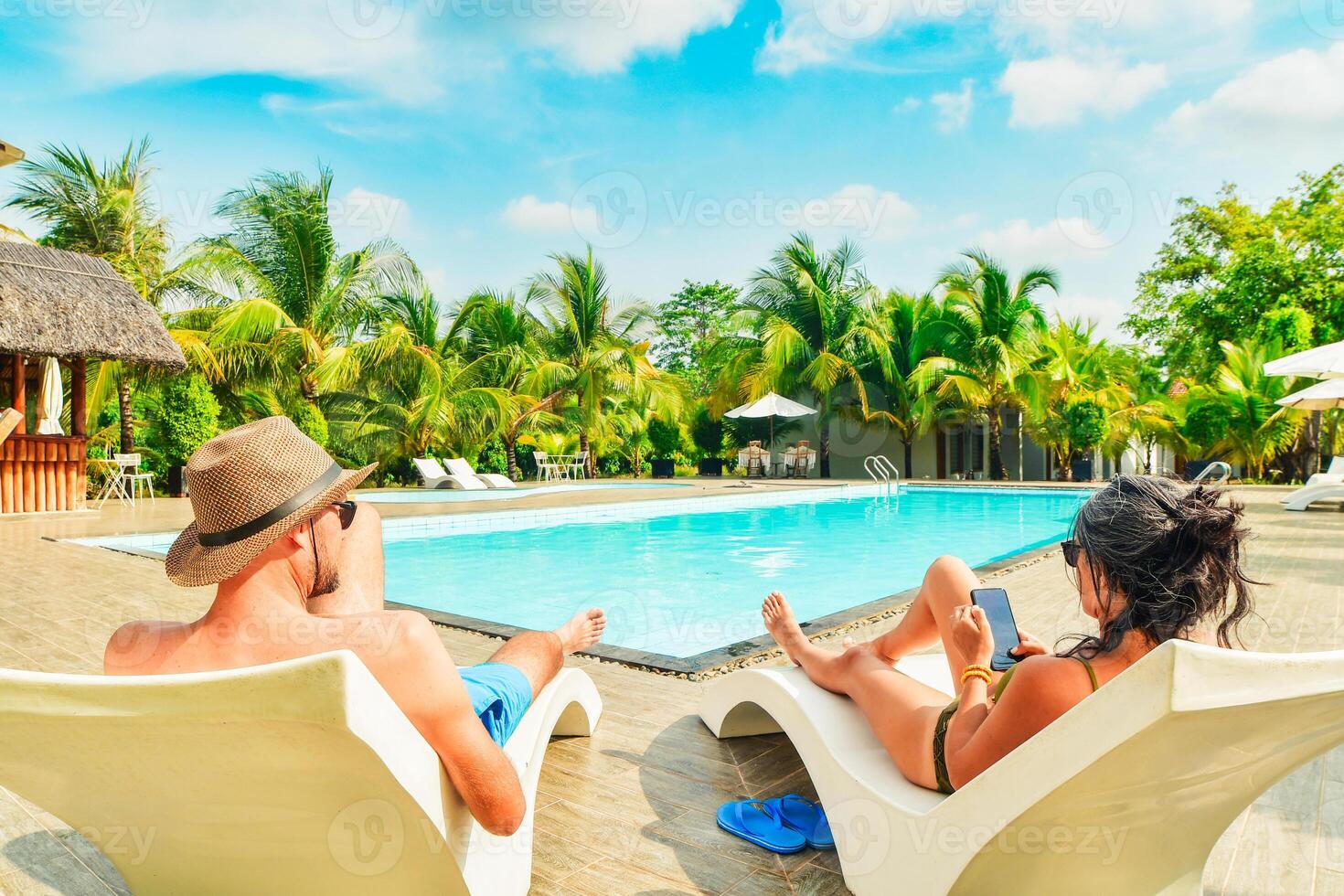 Couple lay on sunbeds enjoy sunbathing relax scroll phone internet on all-inclusive vacation holidays at tropical resort with swimming pool surrounded by coconut palm trees on sunny hot tropical day photo
