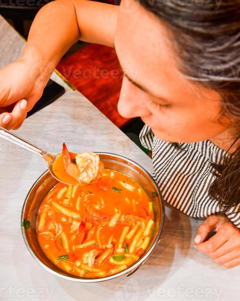 Close up op view caucasian woman tourist sit in asian restaurant try favorite Thai dish Tom Yum hot creamy soup with shrimps in local food restaurant. Thailand vacation food tour concept photo