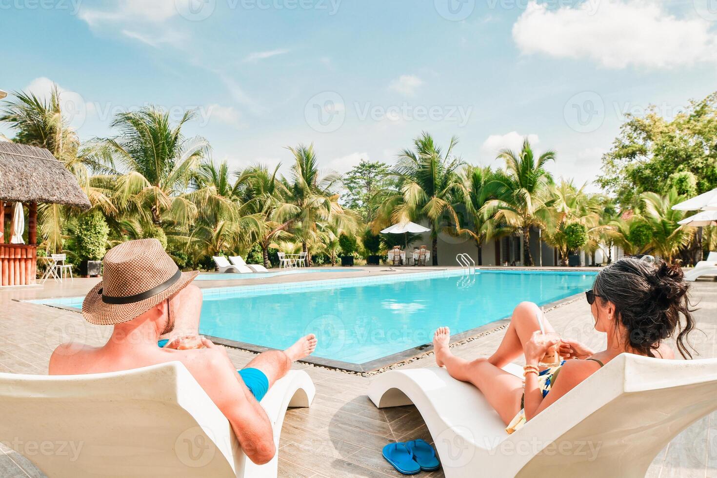 Couple lay on sunbeds enjoy sunbathing drink cold beverage on all-inclusive vacation holidays at tropical resort with swimming pool surrounded by coconut palm trees by coast on sunny hot tropical day photo