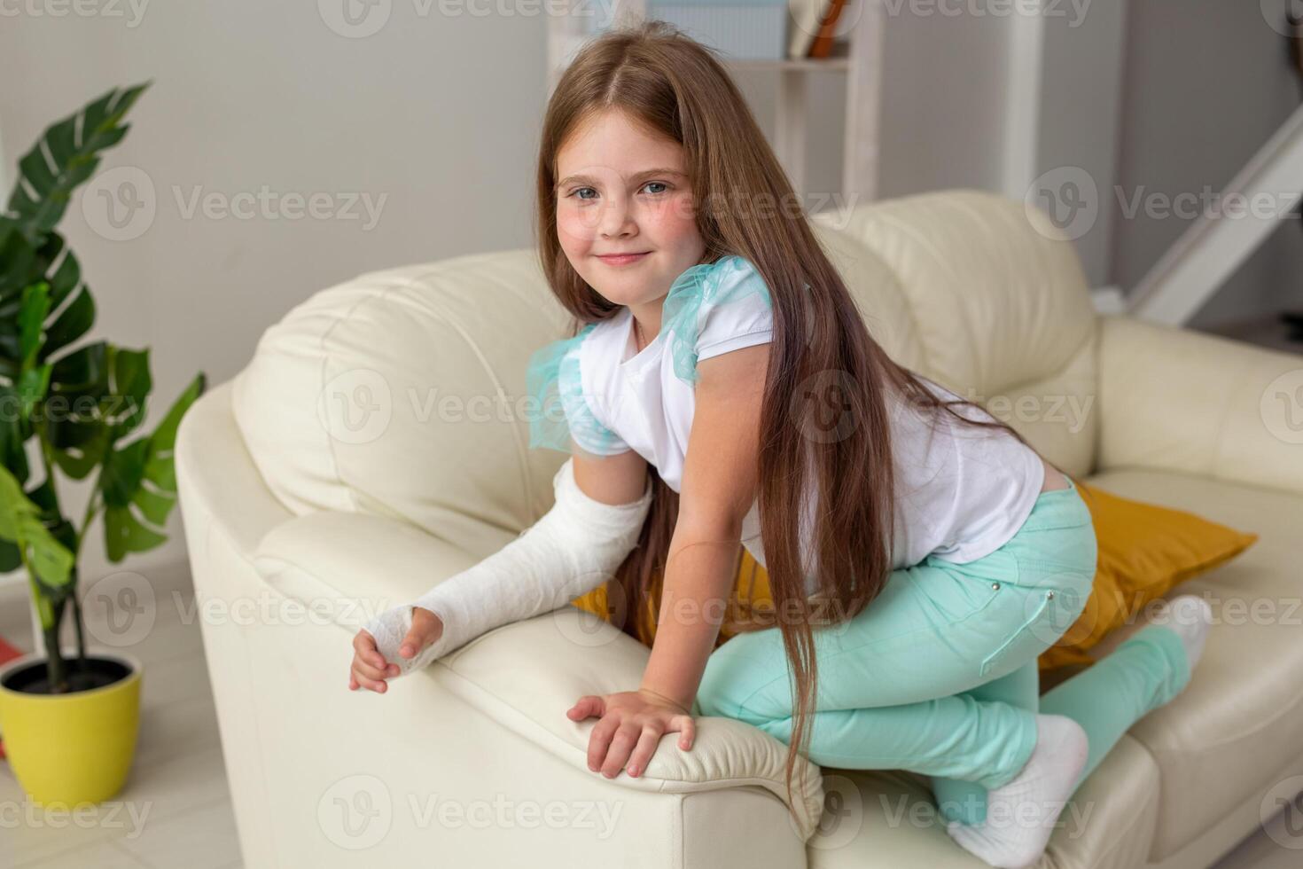 Child with a cast on a broken wrist or arm smiling and having fun on a couch. Positive attitude, recovery and kid concept. photo