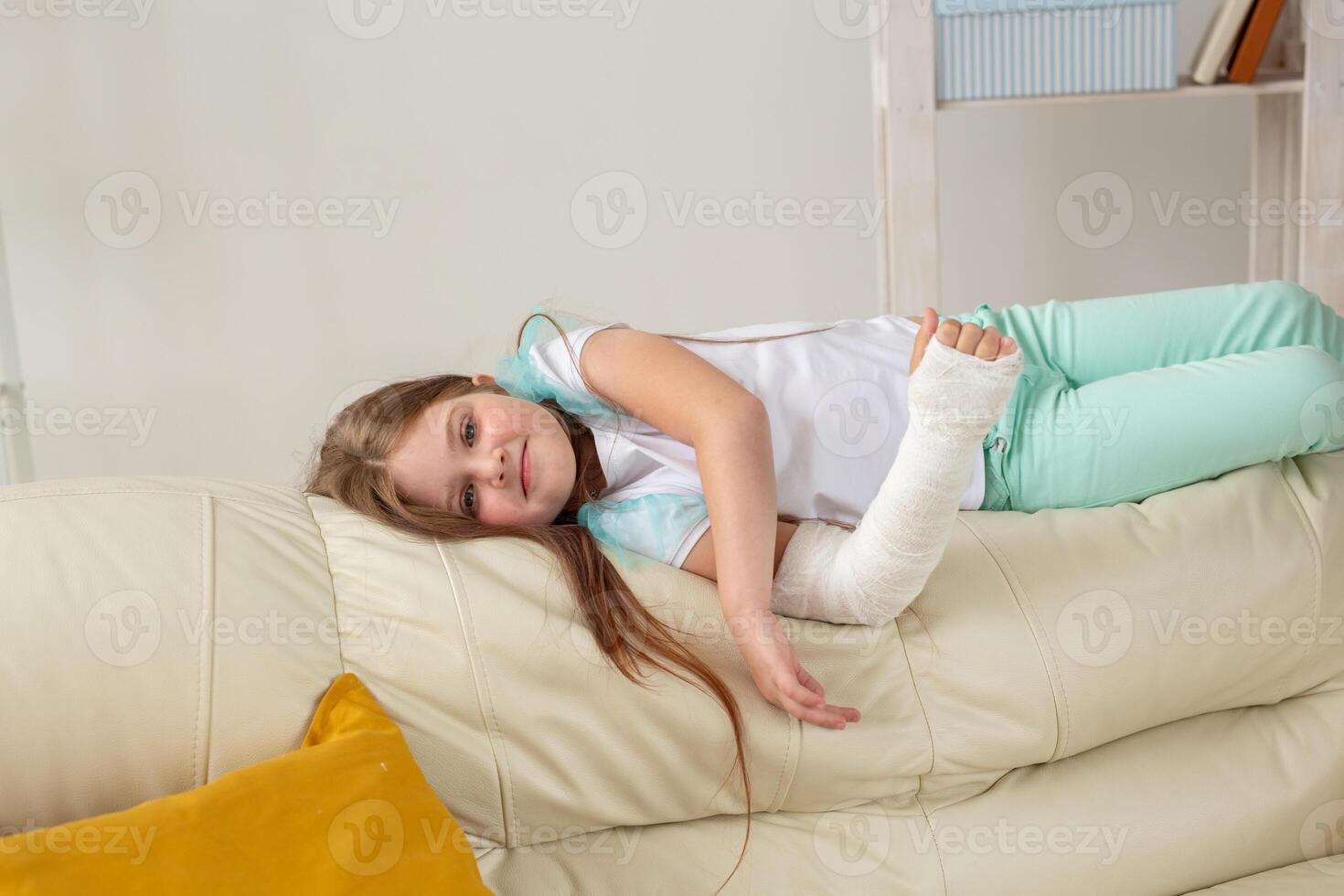 Child with a cast on a broken wrist or arm smiling and having fun on a couch. Positive attitude, recovery and kid concept. photo