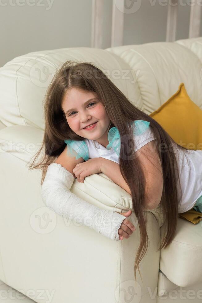 Child with a cast on a broken wrist or arm smiling and having fun on a couch. Positive attitude, recovery and kid concept. photo