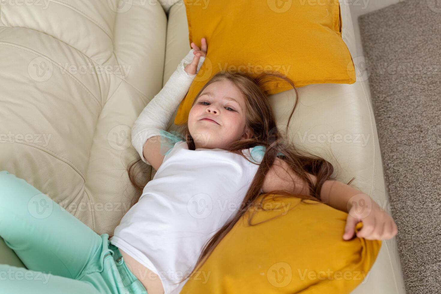 Child with a cast on a broken wrist or arm smiling and having fun on a couch. Positive attitude, recovery and kid concept. photo