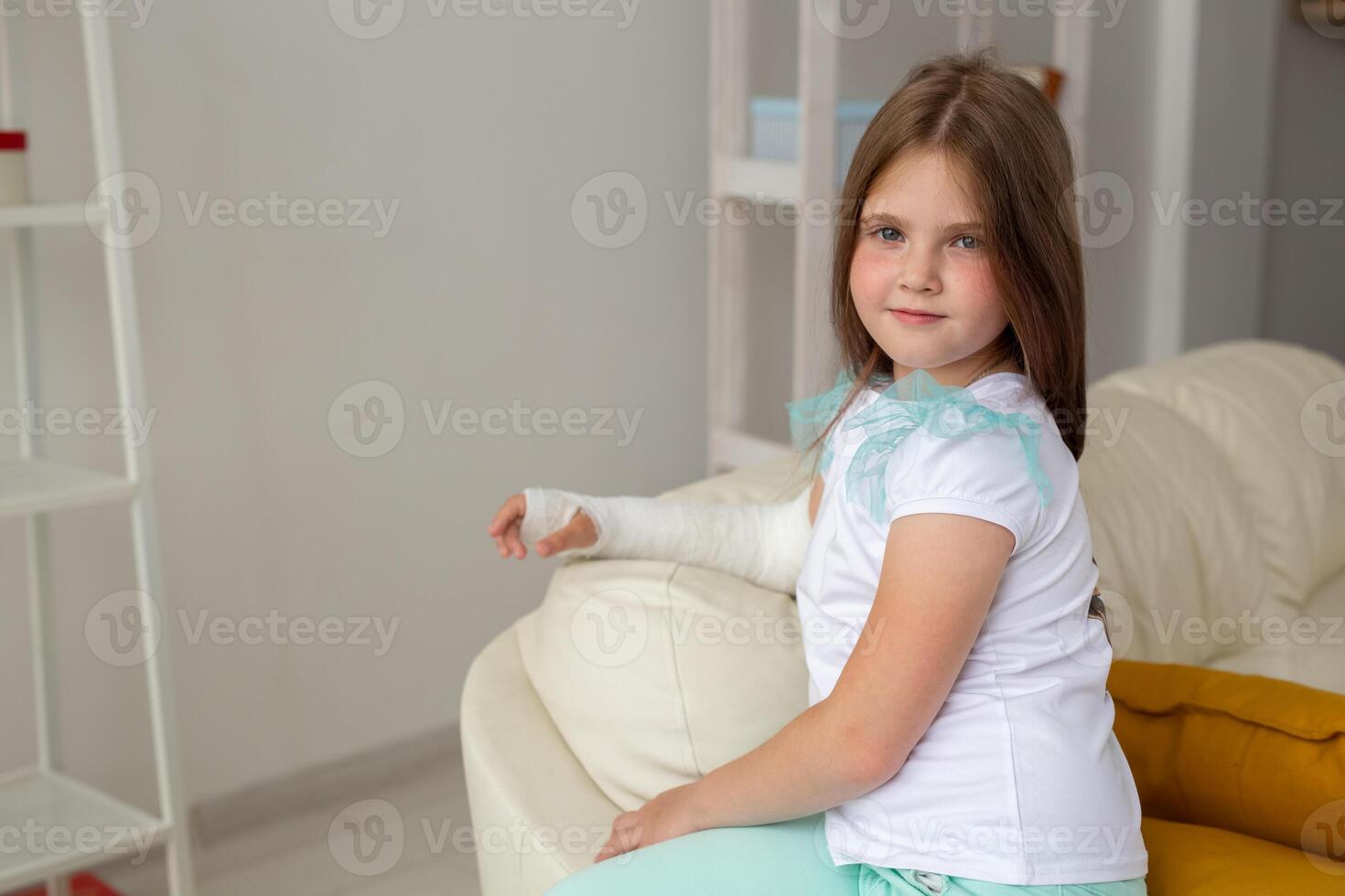 Child with a cast on a broken wrist or arm smiling and having fun on a couch. Positive attitude, recovery and kid concept. photo