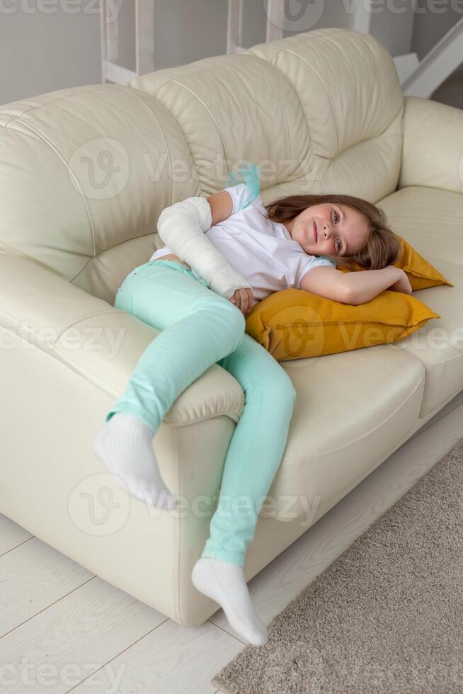Child with a cast on a broken wrist or arm smiling and having fun on a couch. Positive attitude, recovery and kid concept. photo