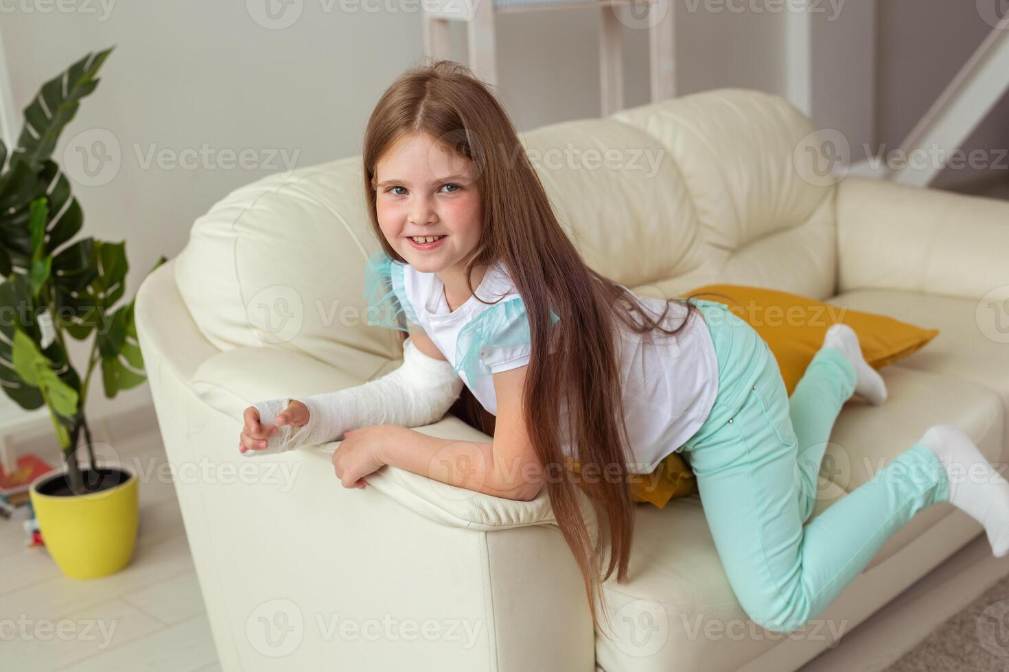 Child with a cast on a broken wrist or arm smiling and having fun on a couch. Positive attitude, recovery and kid concept. photo