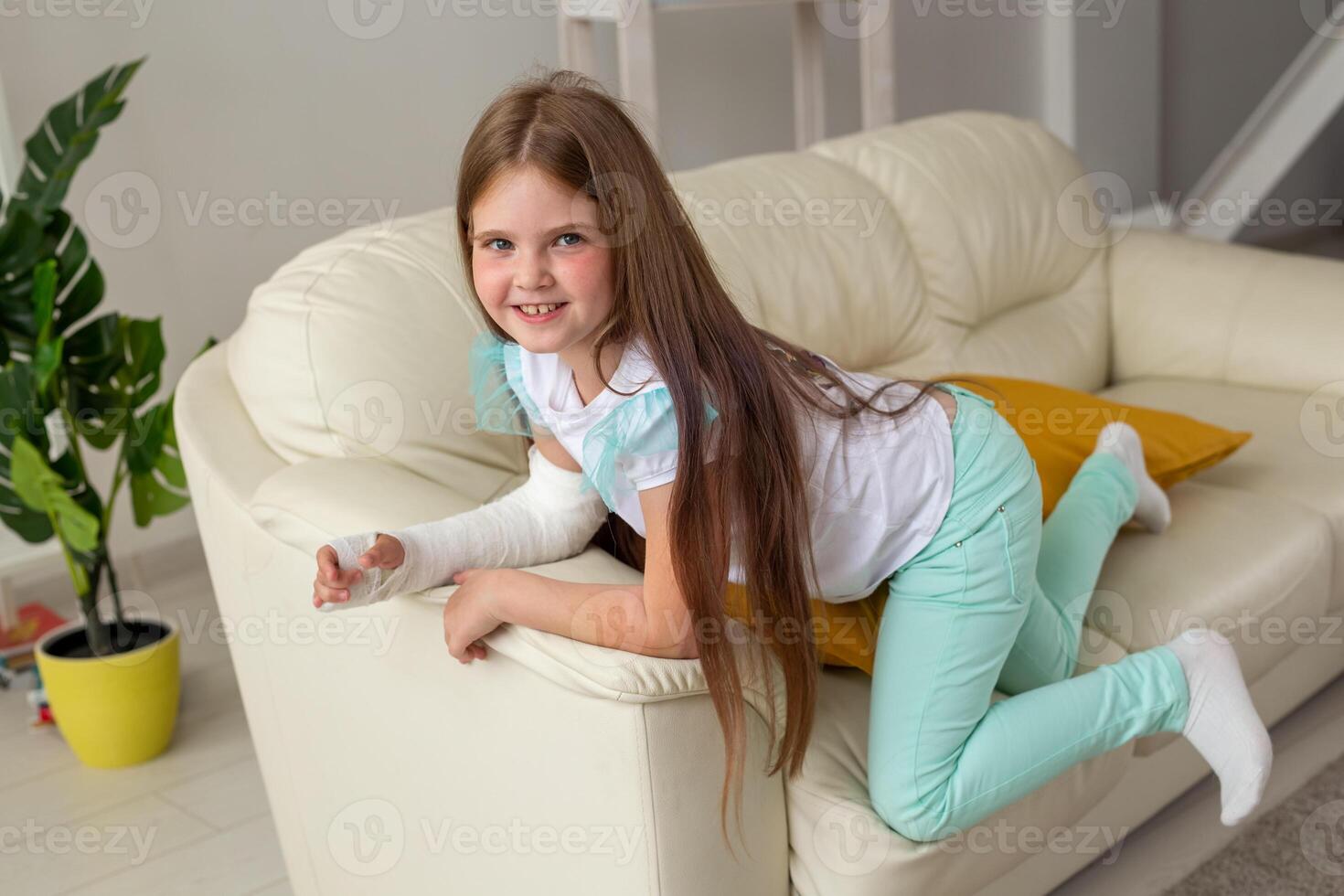 Child with a cast on a broken wrist or arm smiling and having fun on a couch. Positive attitude, recovery and kid concept. photo