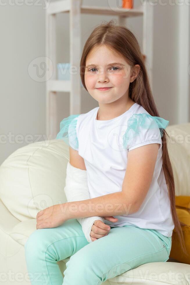 Child with a cast on a broken wrist or arm smiling and having fun on a couch. Positive attitude, recovery and kid concept. photo