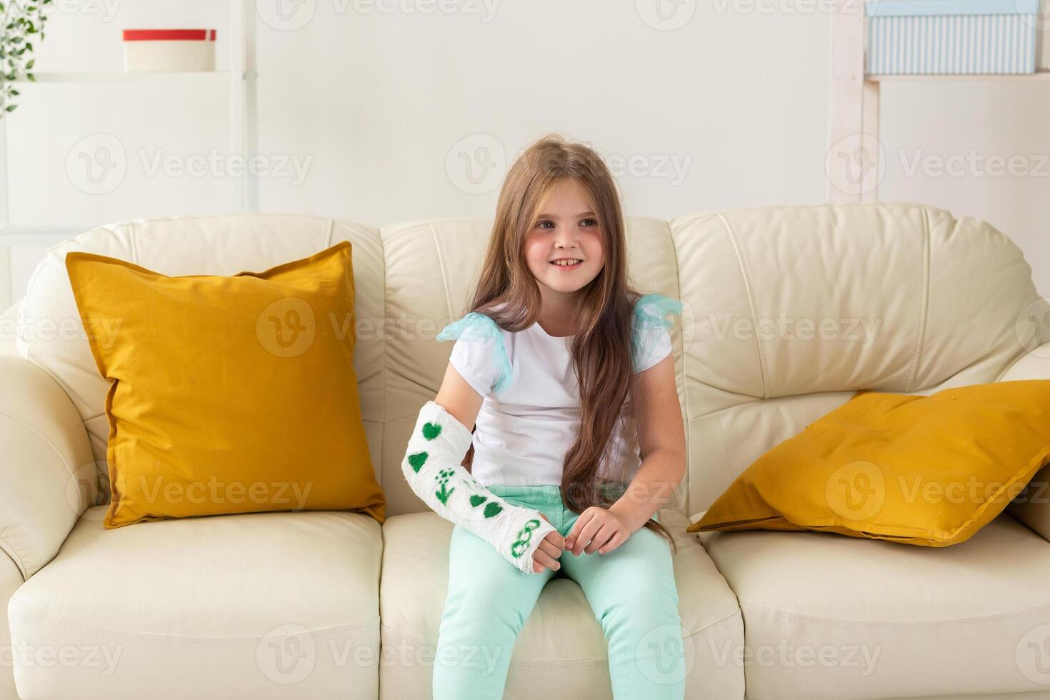 Child with a cast on a broken wrist or arm smiling and having fun on a couch. Positive attitude, recovery and kid concept. photo