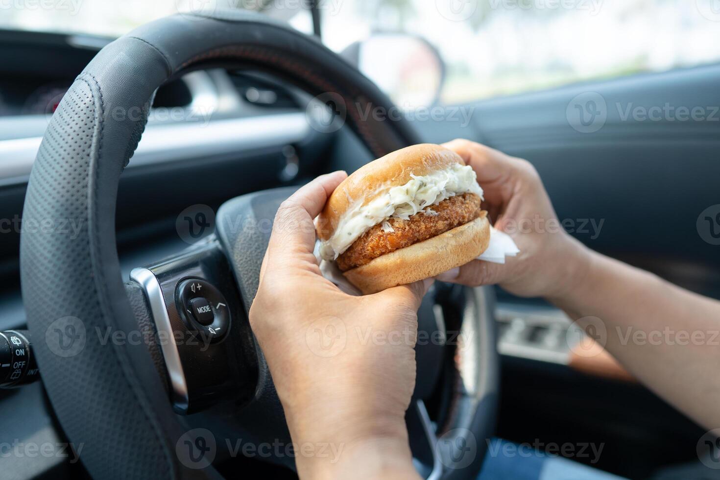 Asian lady holding hamburger to eat in car, dangerous and risk an accident. photo