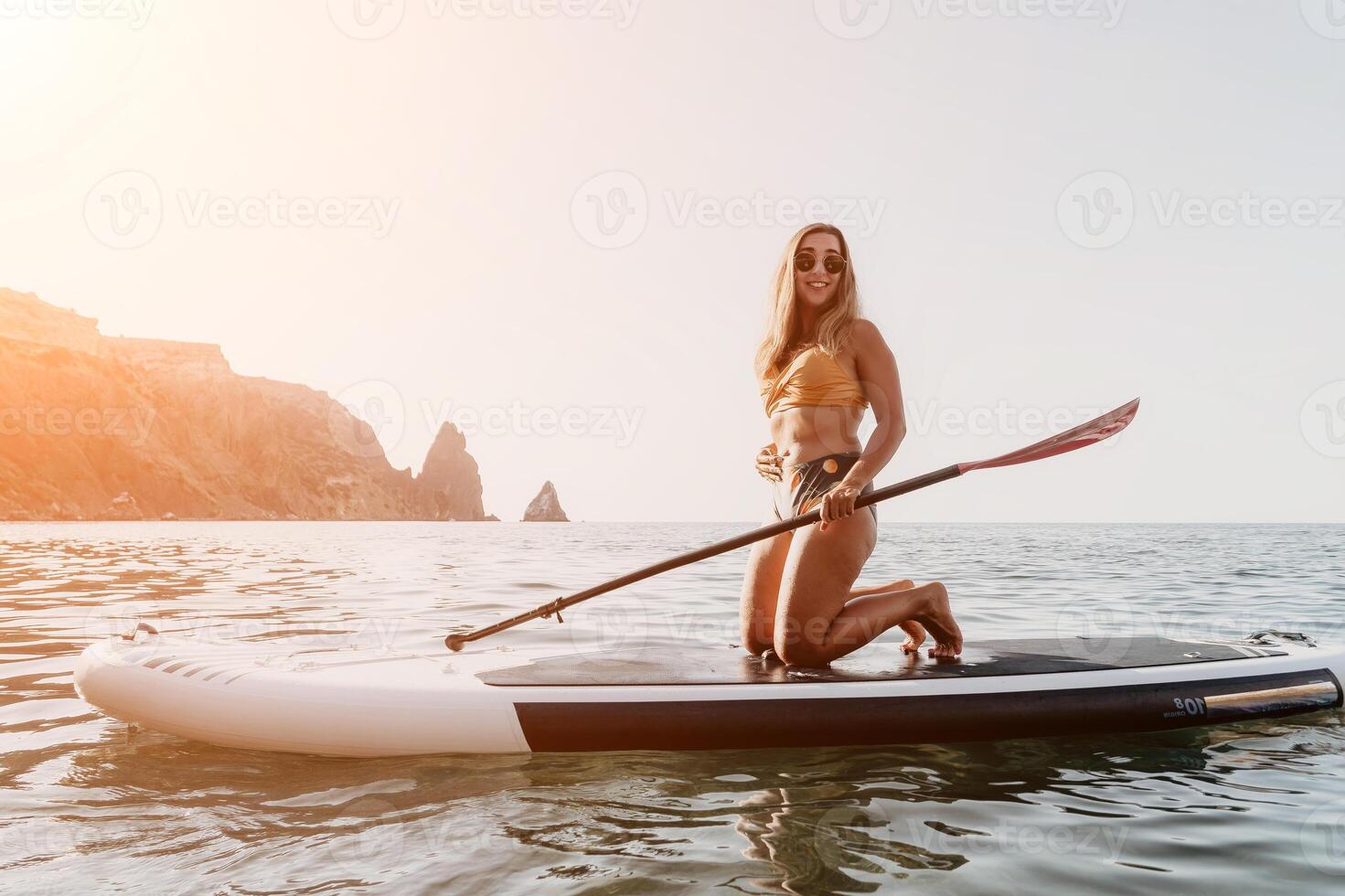 Woman Paddle Boarding Ocean Coastline - Young woman paddling on a stand up paddle board on a calm ocean with rocky cliffs in the background. photo
