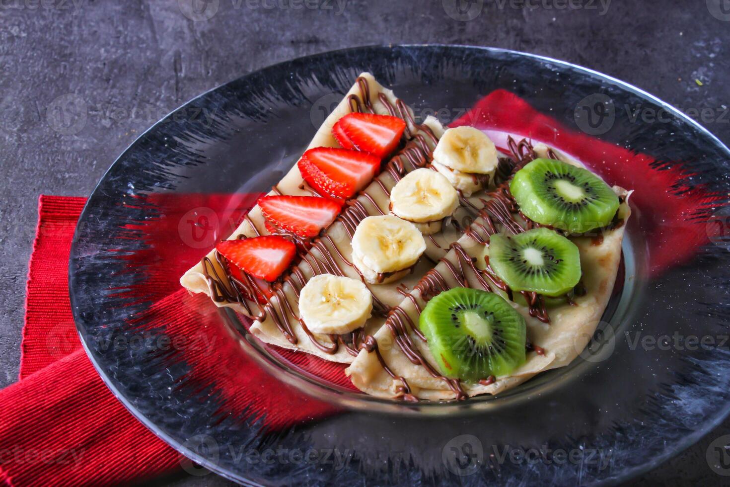 Colorful Fruit Crepe with Chocolate opped with fresh strawberries, bananas, and kiwi slices, with rich chocolate sauce served in plate isolated on table side view of indulgent dessert photo