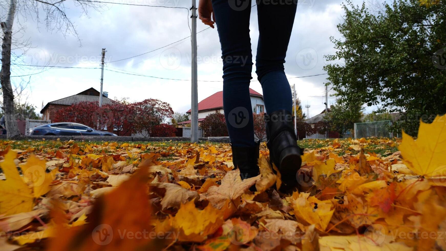 Legs of young woman stepping on yellow foliage at park. Female feet in boots going on fallen maple leaves at parkland. Girl walking at parkland at autumn season on cloudy day. Close up Slow motion photo