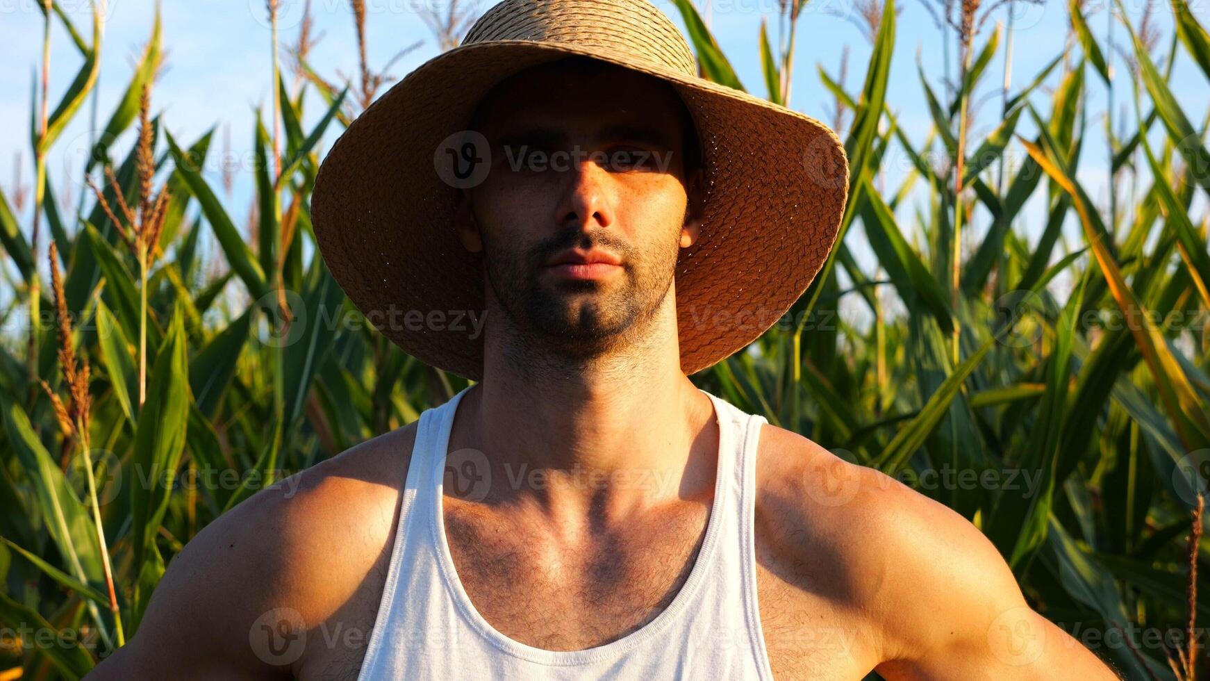 Serious male farmer looks into camera standing near corn field. Portrait of young handsome agronomist in straw hat with maize meadow at background. Concept of agricultural business. Close up photo