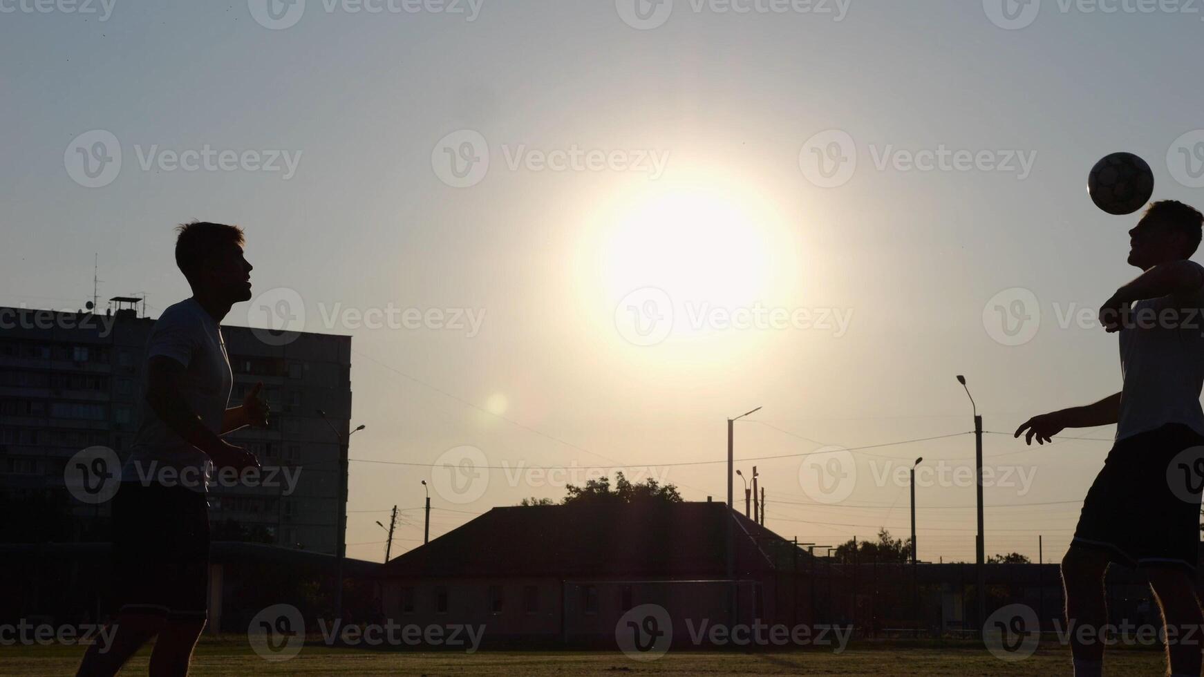 Silhouette of professional footballers kicking soccer ball with their heads on stadium at sunset. Two sportsmen showing tricks with ball while passing it to each other at field. Freestyle football photo