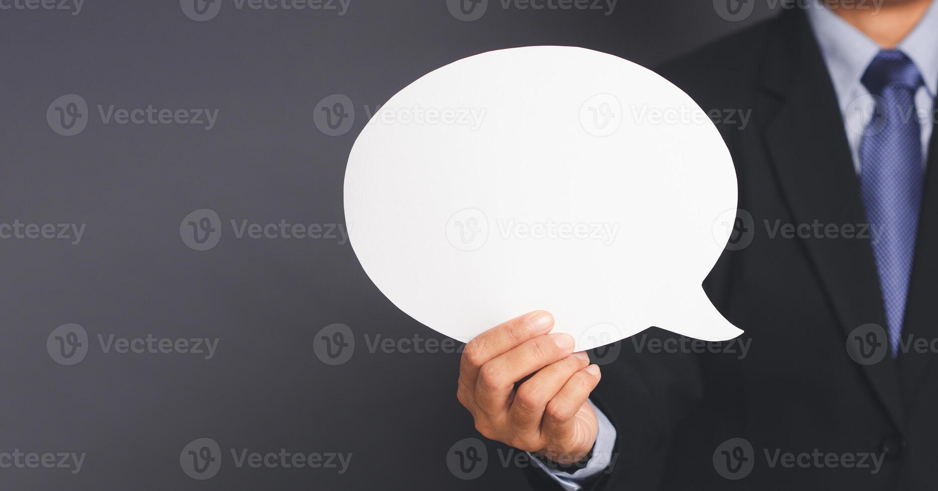 Business and speech bubble concept. Businessman in suit hand holding an empty speech bubble paper while standing on gray background in the studio. Space for text. Close-up photo