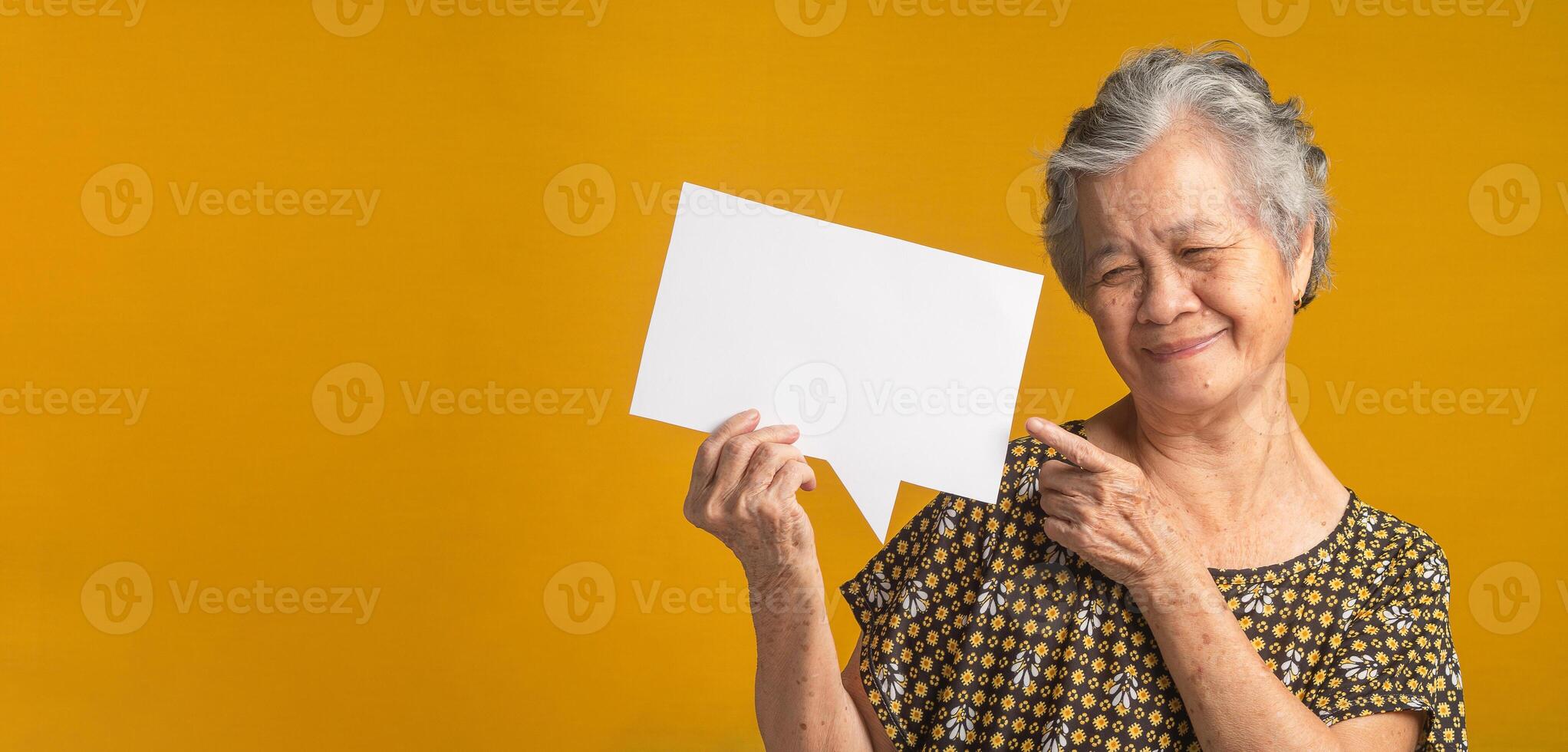 Concept of a paper speech bubble. An elderly Asian woman with short gray hair holding looking and pointing at a blank paper with a smile while standing on a yellow background in the studio photo
