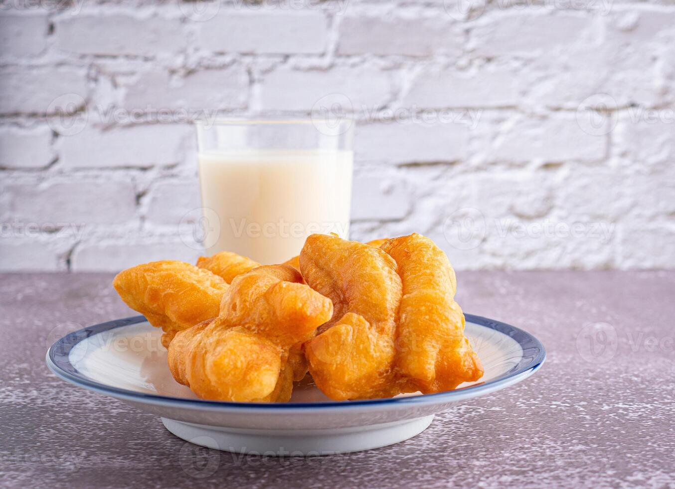 Close-up of deep-fried dough sticks or Chinese doughnut sticks on white plate and a glass of soybean milk on gray stone background with white brick wall. Space for text photo