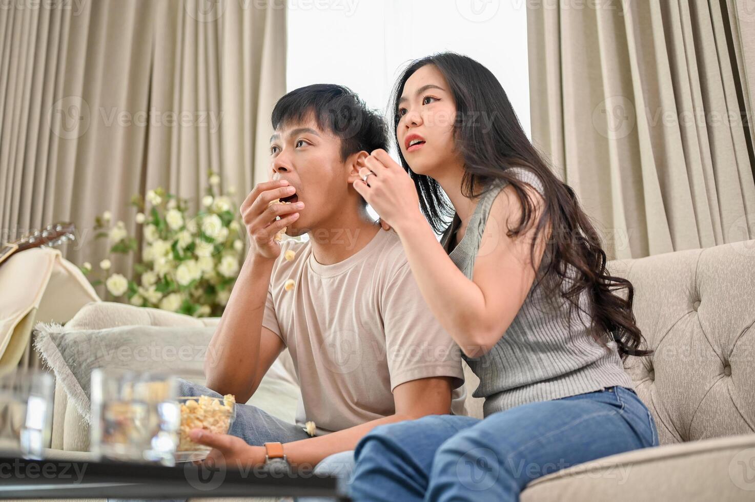 A shocked young Asian couple is watching an exciting TV show or movie on a sofa together at home. photo