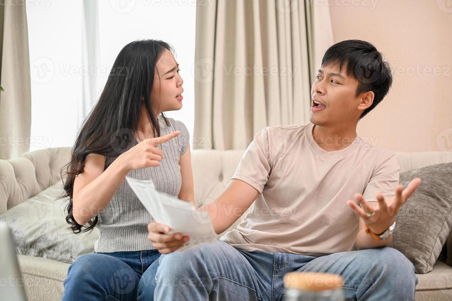 A resentful, angry Asian wife and husband are arguing about family household invoices on sofa. photo