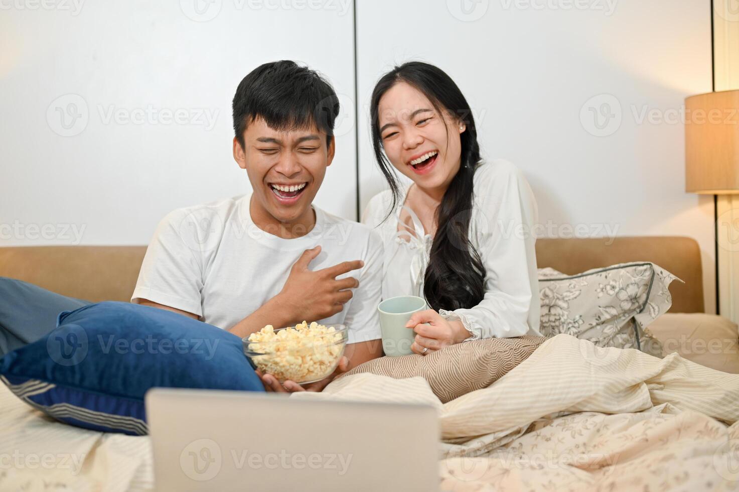 A cheerful Asian couple is laughing, enjoying watching a movie on a laptop in bed together. photo