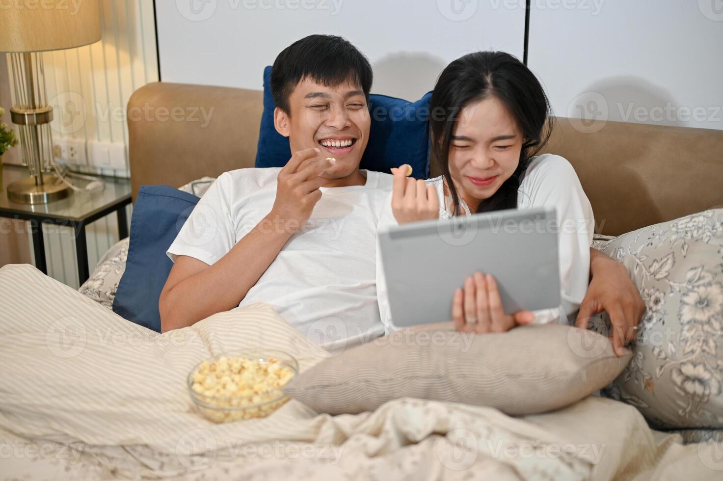 A happy Asian couple are enjoying watching a movie on a tablet and eating popcorn in bed together. photo