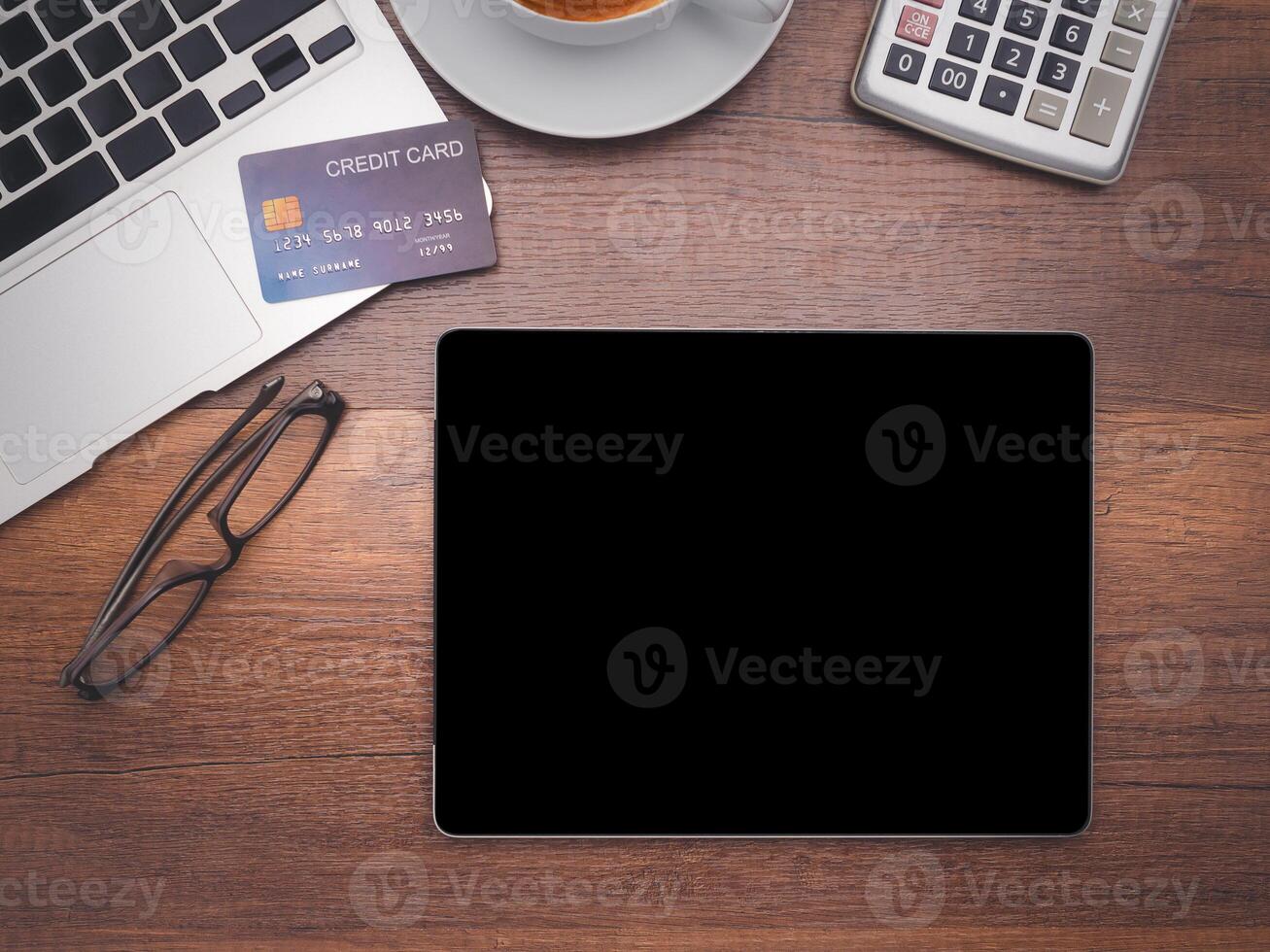 Top view of a tablet with a laptop, credit card, glasses, a coffee cup, and calculator on wooden table in the office. Flat lay, space for text. Working desk table concept photo