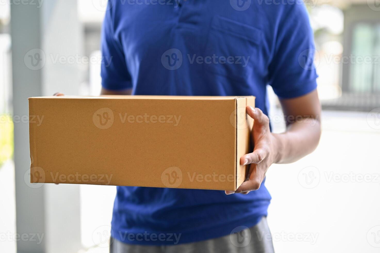 A cropped shot of a delivery man in a blue shirt holding a cardboard box in front of the door. photo
