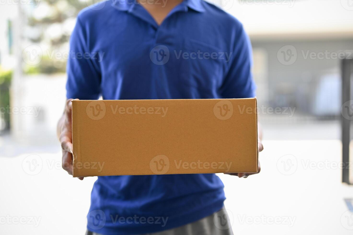 A cropped shot of a delivery man in a blue shirt holding a cardboard box in front of the door. photo