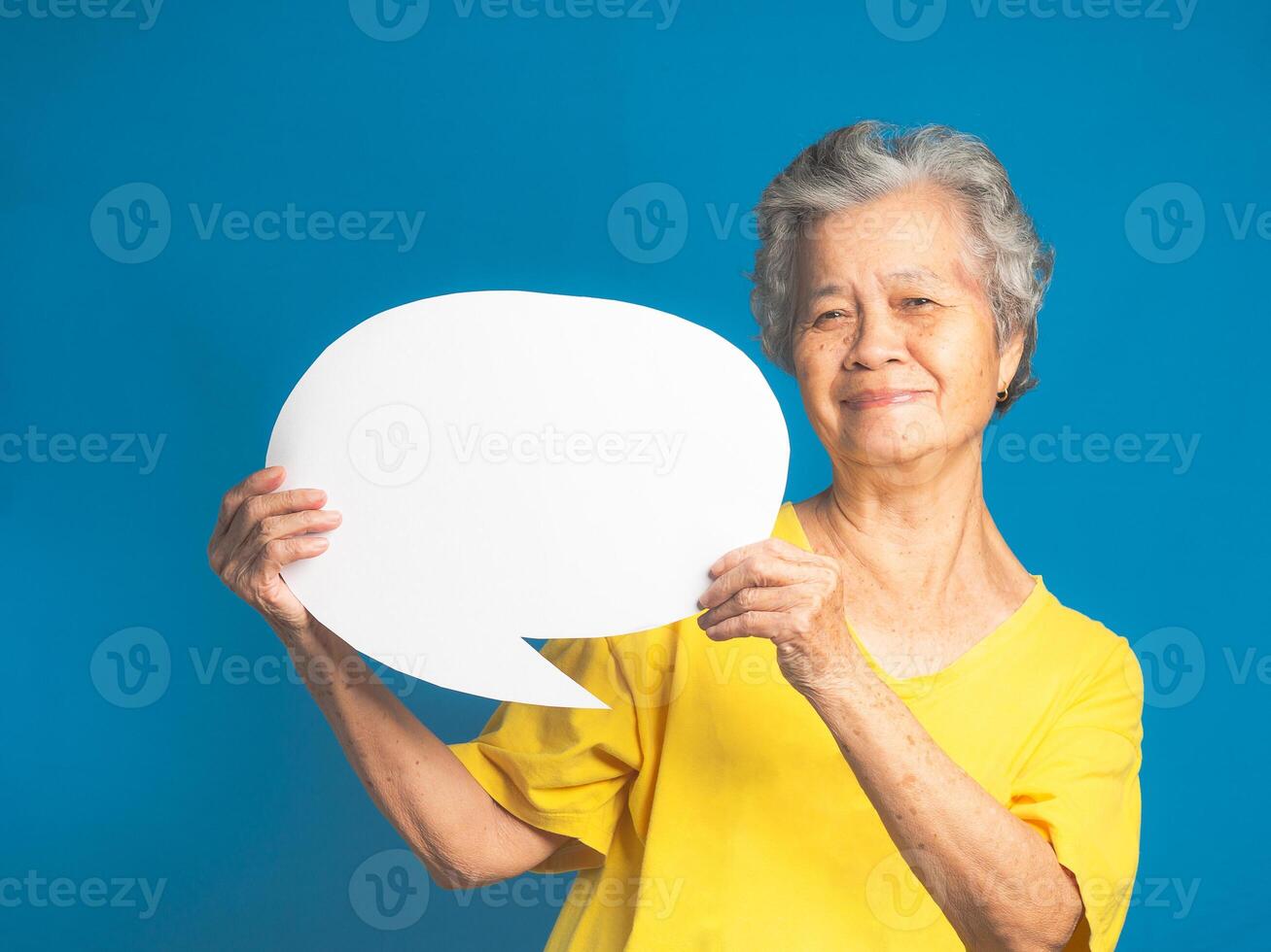 Concept of a speech bubble. Happy senior woman with short gray hair holding a blank paper and looking at the camera with a smile while standing on a blue background in the studio photo