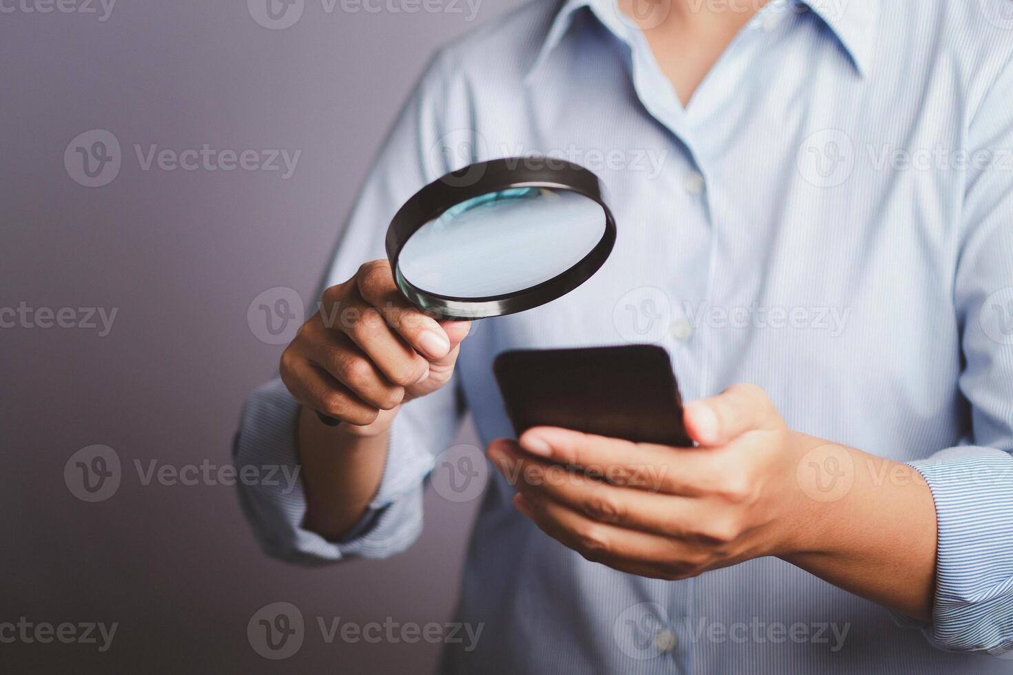 Close-up of the hand of a businesswoman wearing a blue shirt holding a magnifying glass looking at her phone while standing in a gray background in the studio photo