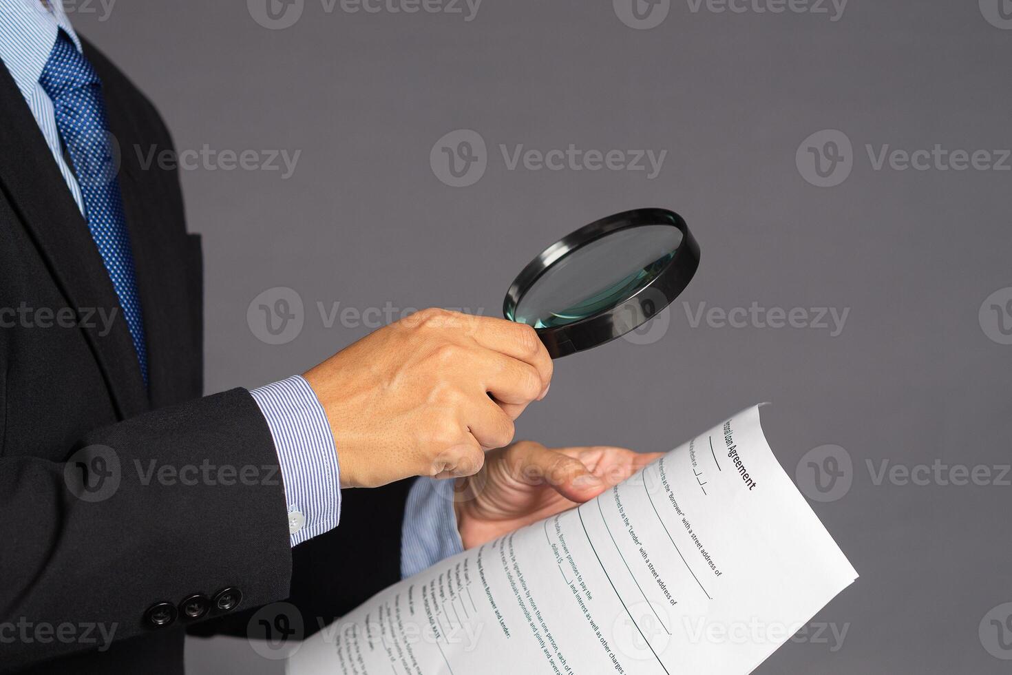 Businessman in a suit holding a magnifying glass and reading a document while standing over a gray background with copy space for text. Business and healthcare concept photo