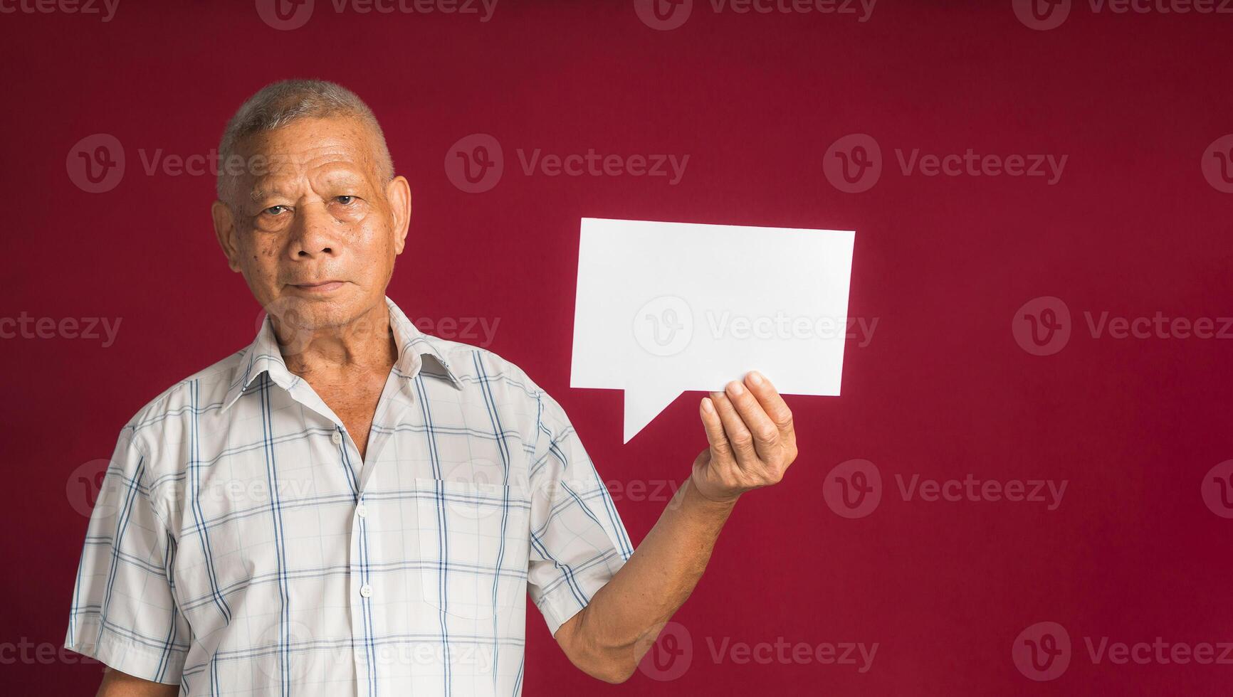 Concept of a speech bubble. Happy senior man with short gray hair holding a blank paper speech bubble and looking at the camera with a smile while standing on a red background in the studio photo