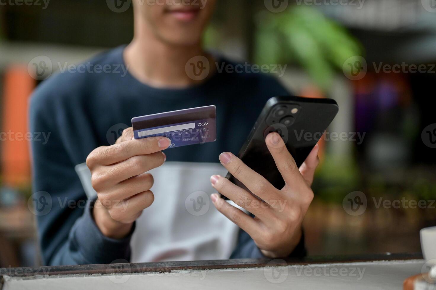 A cropped image of a positive young Asian man holding a smartphone and a credit card over a table. photo