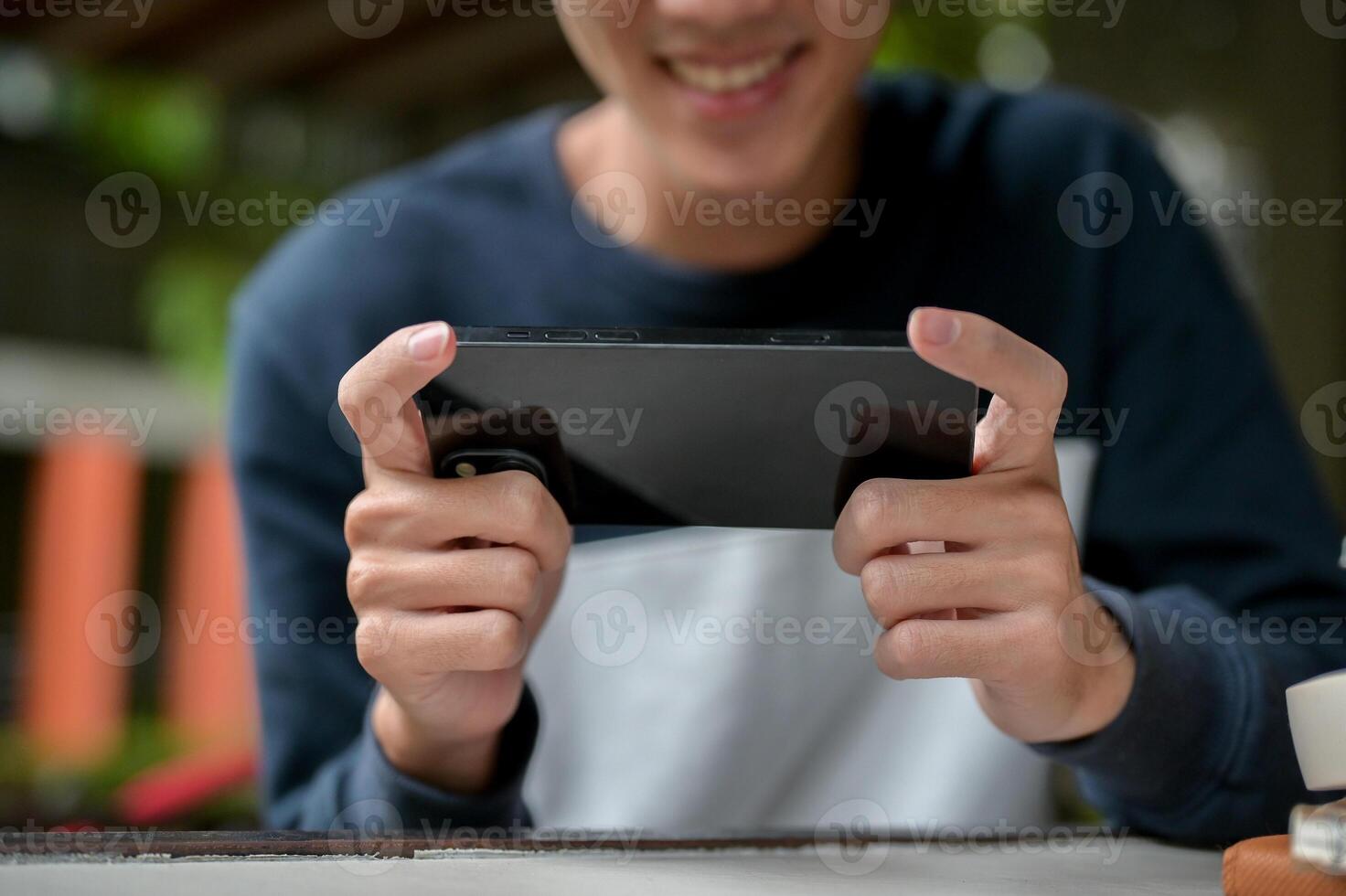 A cropped image of a joyful young Asian man happily playing a mobile game on his smartphone. photo