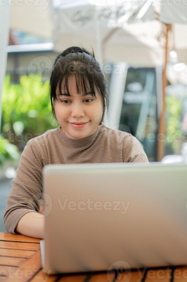 A positive Asian woman working remotely at a cafe in the city, using her laptop. photo