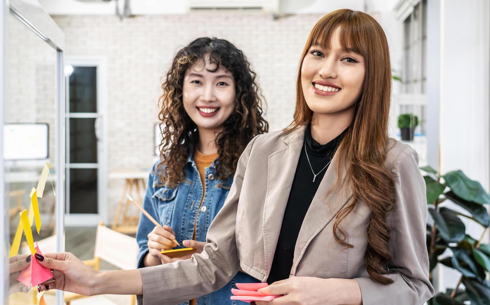 Asian creative business women are writing note on the text sheet after thinking new idea and planning project with colleague, Two employee brainstorming idea on glass wall and sticky photo