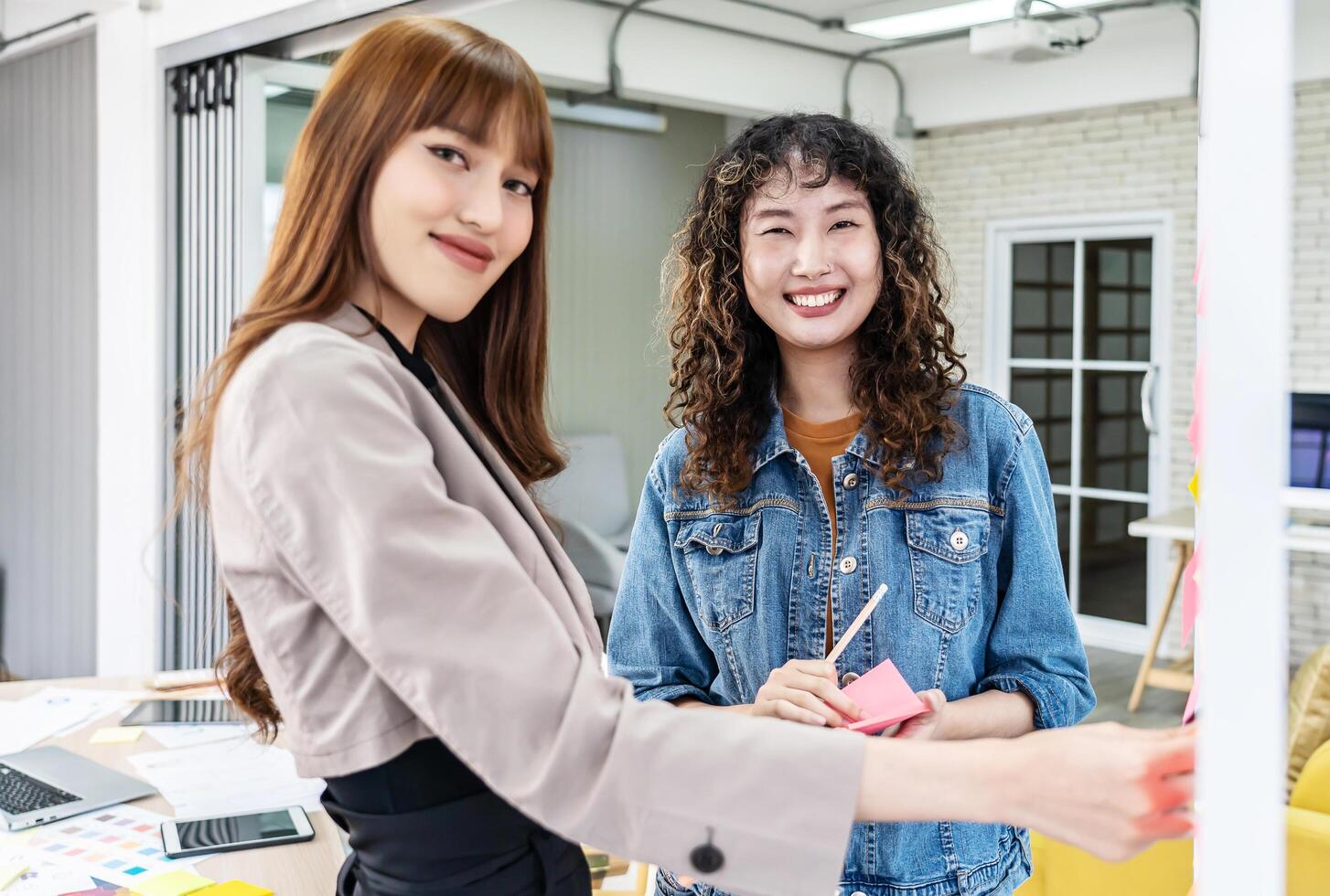 Asian creative business women are writing note on the text sheet after thinking new idea and planning project with colleague, Two employee brainstorming idea on glass wall and sticky photo