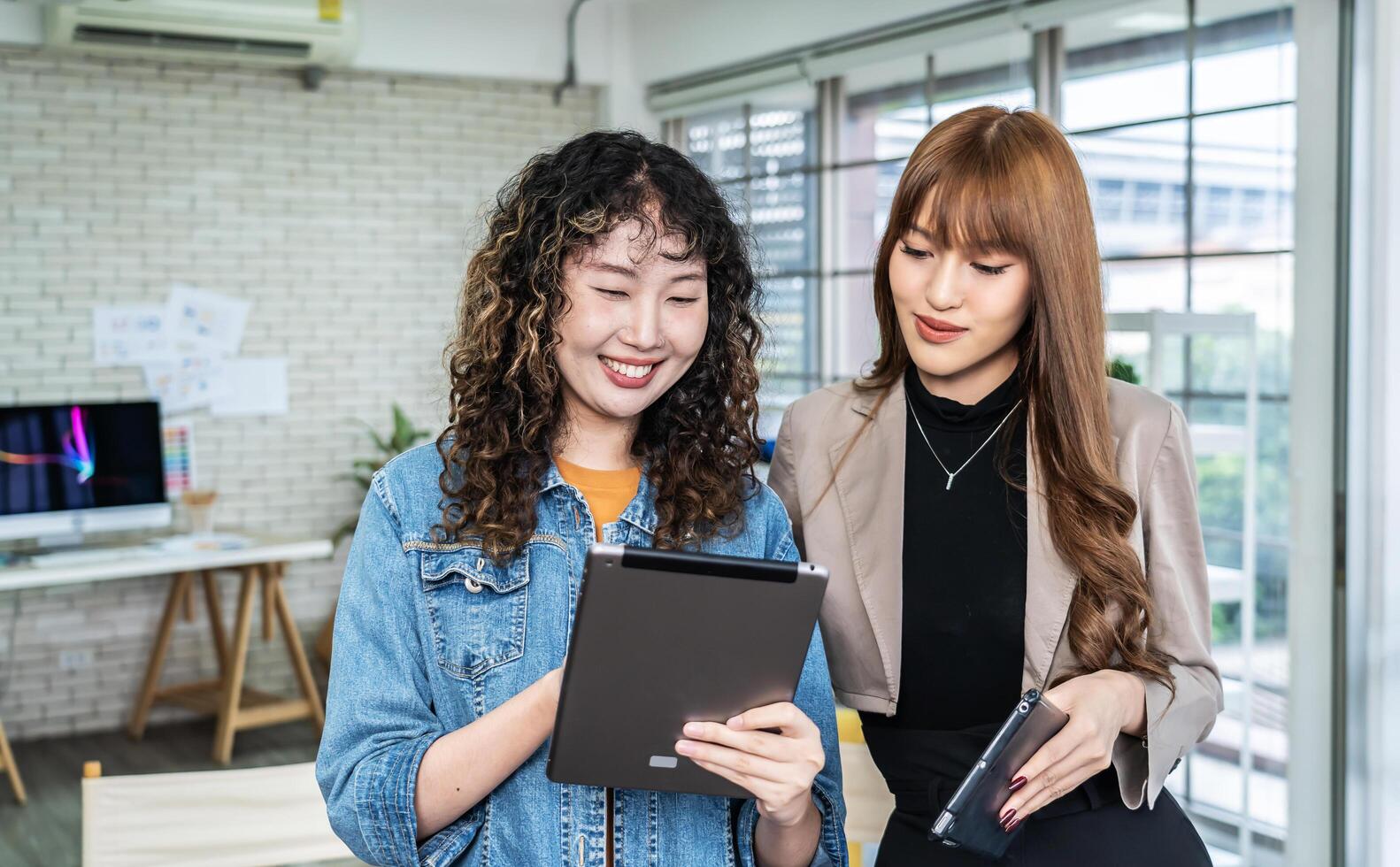 Portrait of cheerful two asian business woman standing in office room discussing collaborative project by using tablet. specifies details of task, talk to mentor at workspace.Teamwork photo