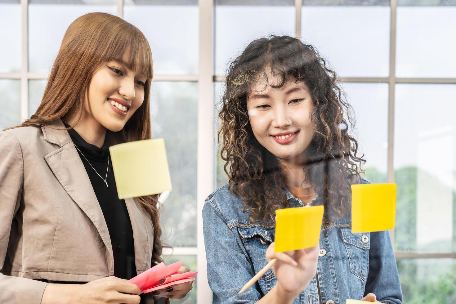 Asian creative business women are writing note on the text sheet after thinking new idea and planning project with colleague, Two employee brainstorming idea on glass wall and sticky photo