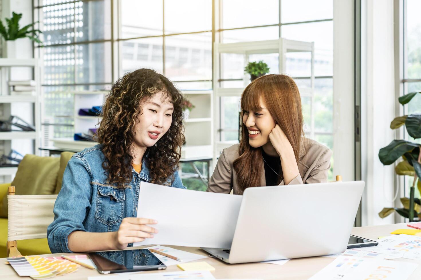 Two business young asian woman working in computer laptop together in office with reports, colleagues discuss financial data analyzing documents marketing project result talk business conversation photo