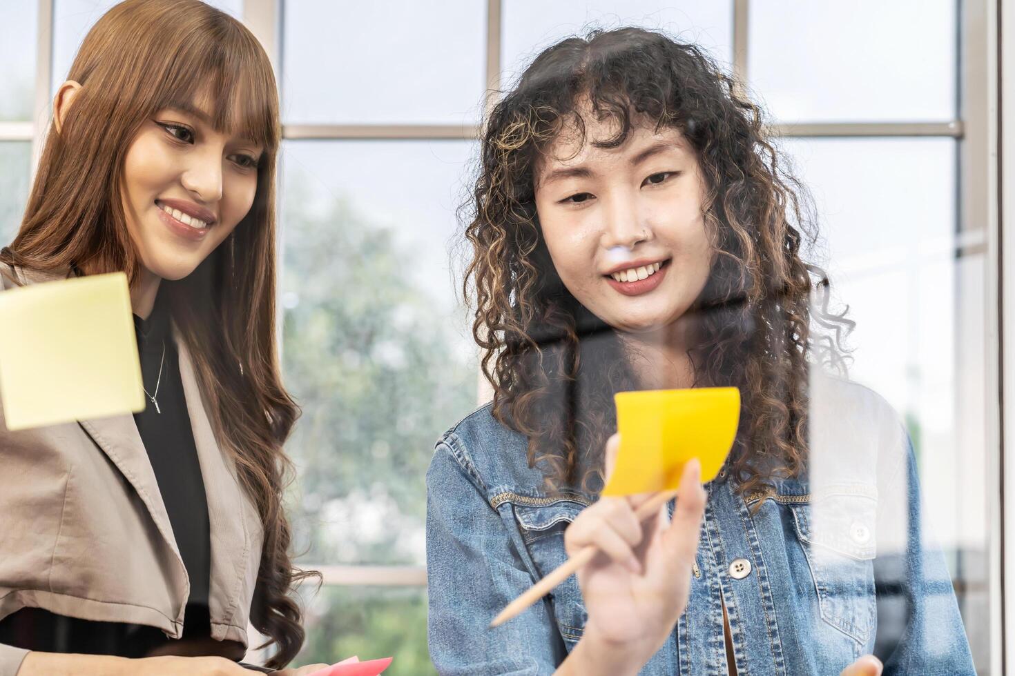 Asian creative business women are writing note on the text sheet after thinking new idea and planning project with colleague, Two employee brainstorming idea on glass wall and sticky photo